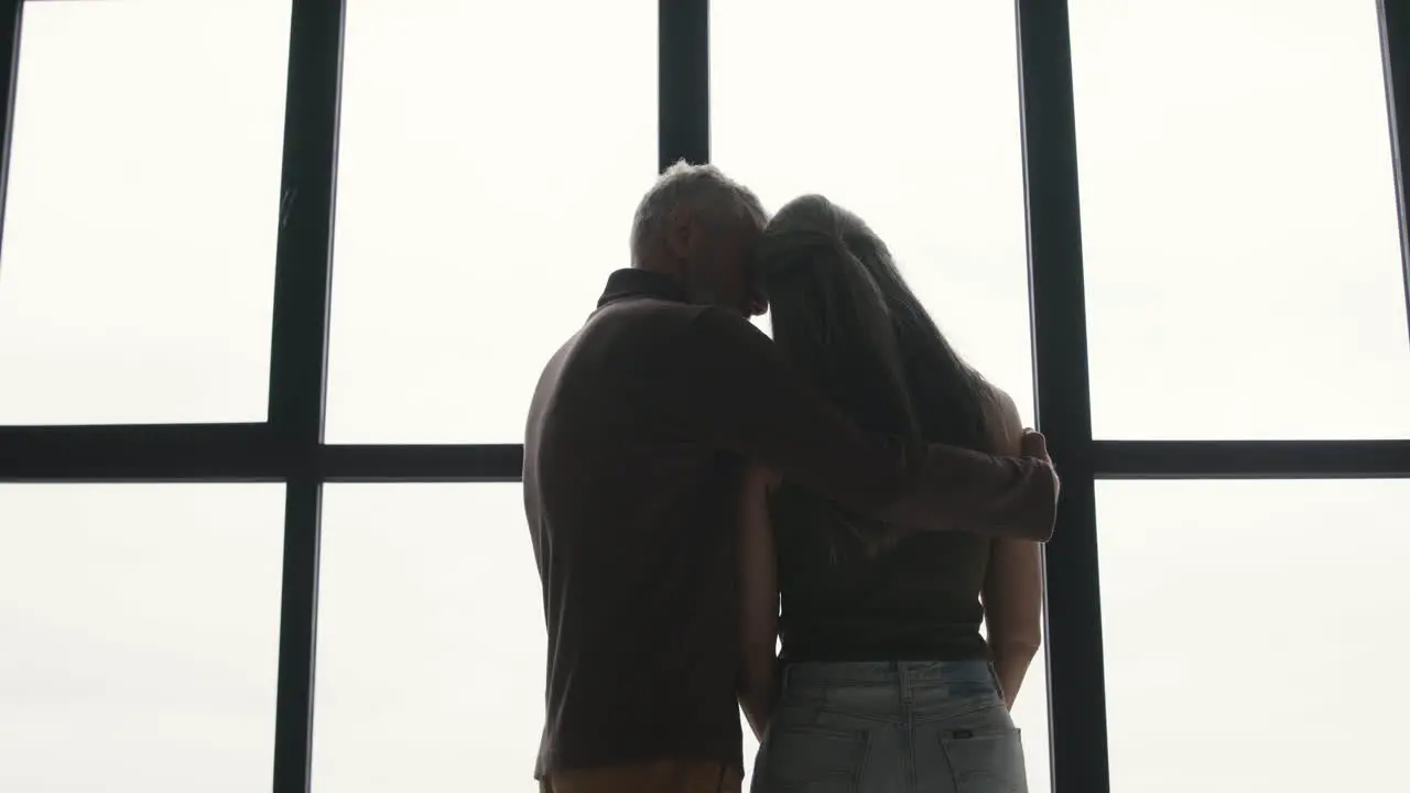 Vista Trasera De Una Triste Mujer De Mediana Edad Con El Pelo Largo Y Gris Parado En La Ventana Mientras Su Marido Intenta Consolarla