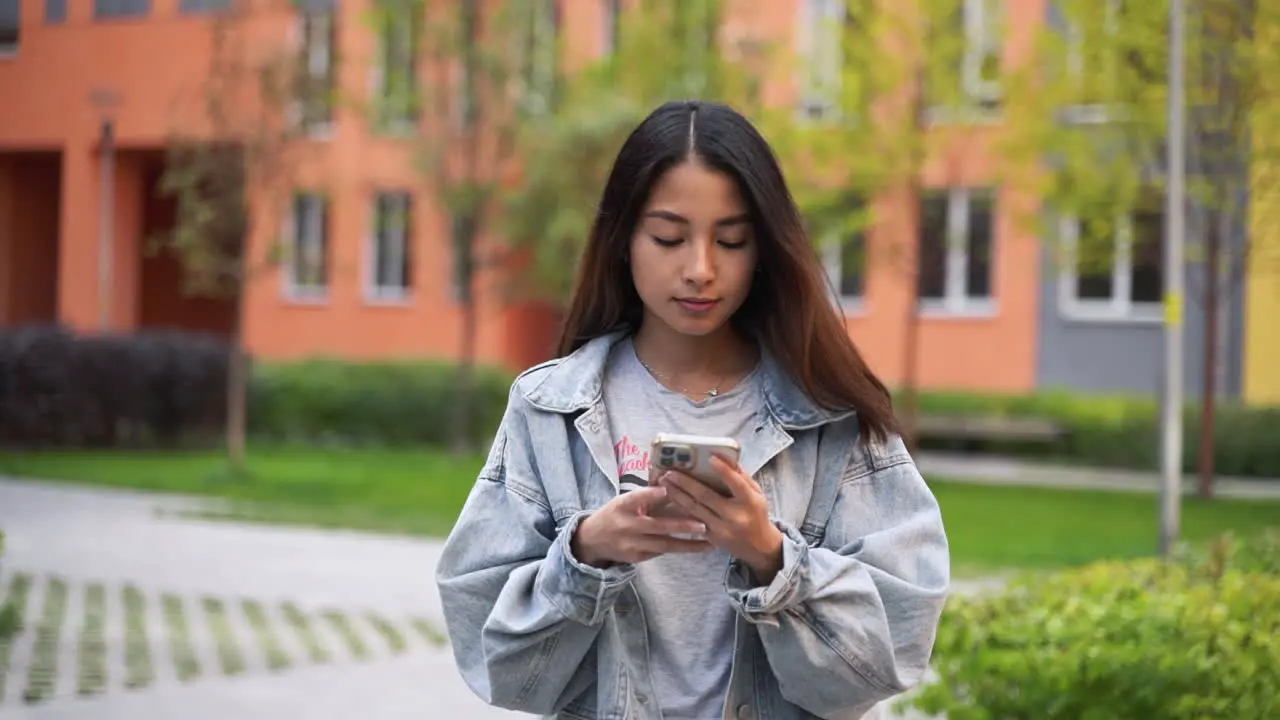 Hermosa Joven Japonesa Enviando Mensajes De Texto En El Teléfono Móvil Mientras Camina Hacia La Cámara Al Aire Libre