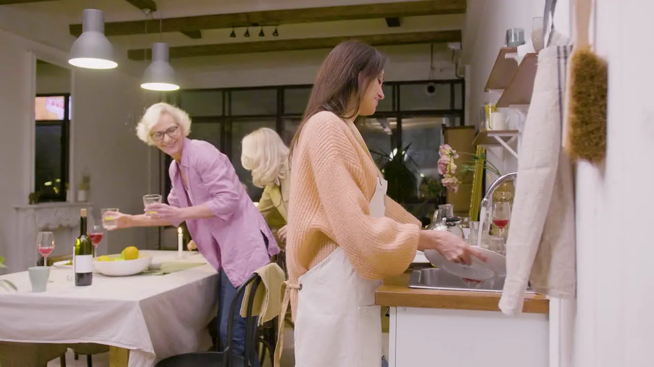 Vista Lateral De Una Mujer Lavando Los Platos De La Cena Familiar En El Fregadero De La Cocina Mientras Dos Mujeres Maduras Quitan Los Platos De La Mesa