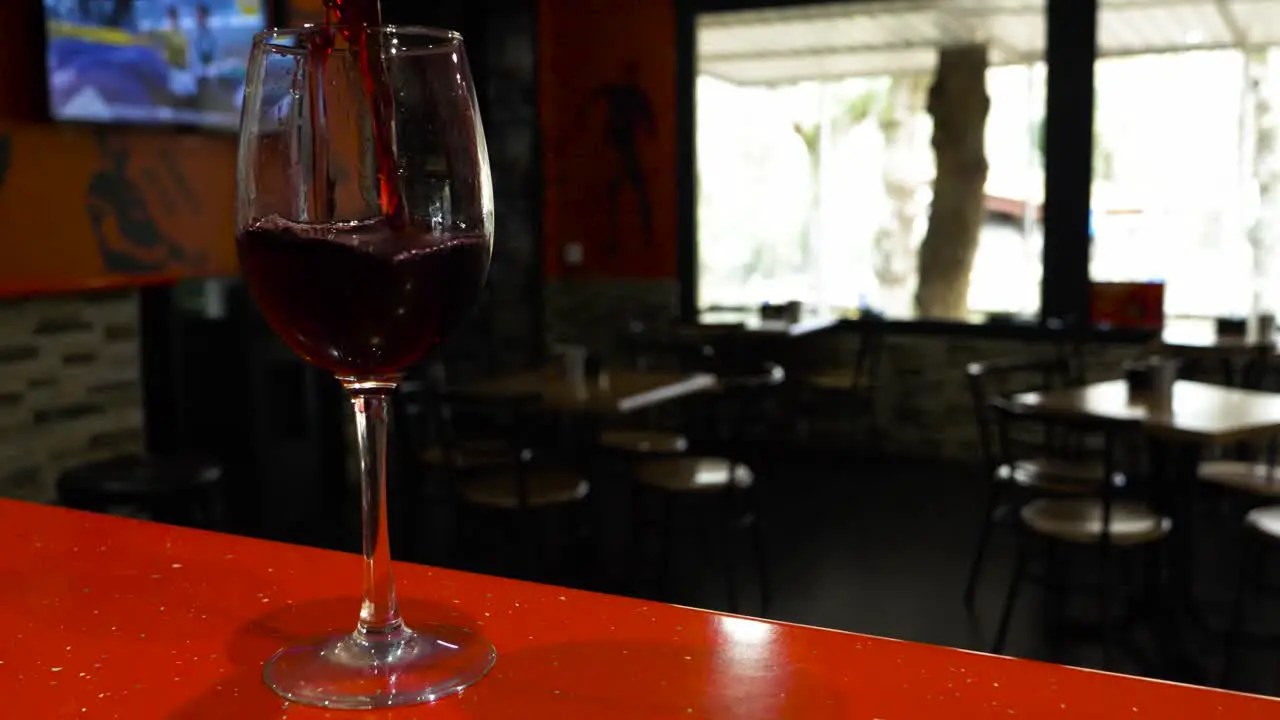 Cooled empty wine glass is filled with red liquid as tv plays in blurred background