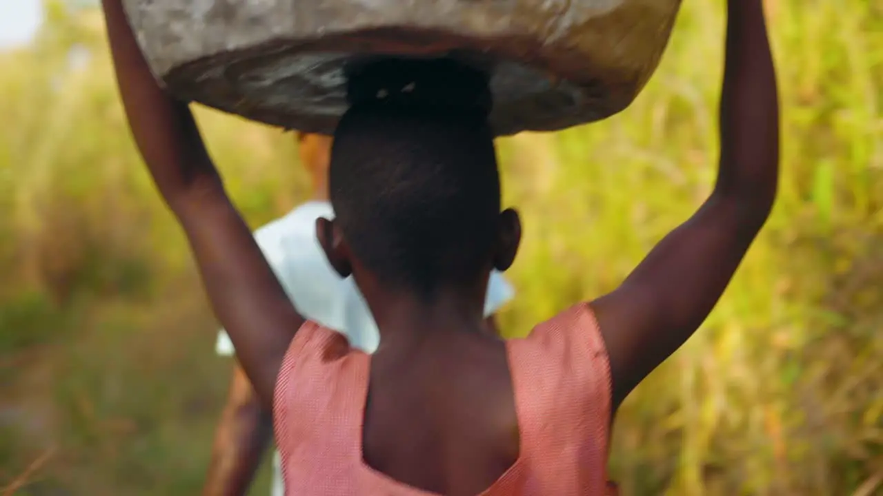 Group of local young African women and kids are carrying big heavy buckets of drinking water and bringing the water on their heads towards the village walking in bush field close shot