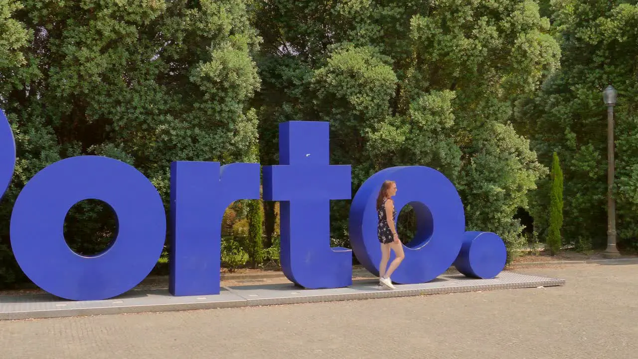 Young tourist woman walking left to right in front of a large blue sign reading "Porto" establishing the cities name