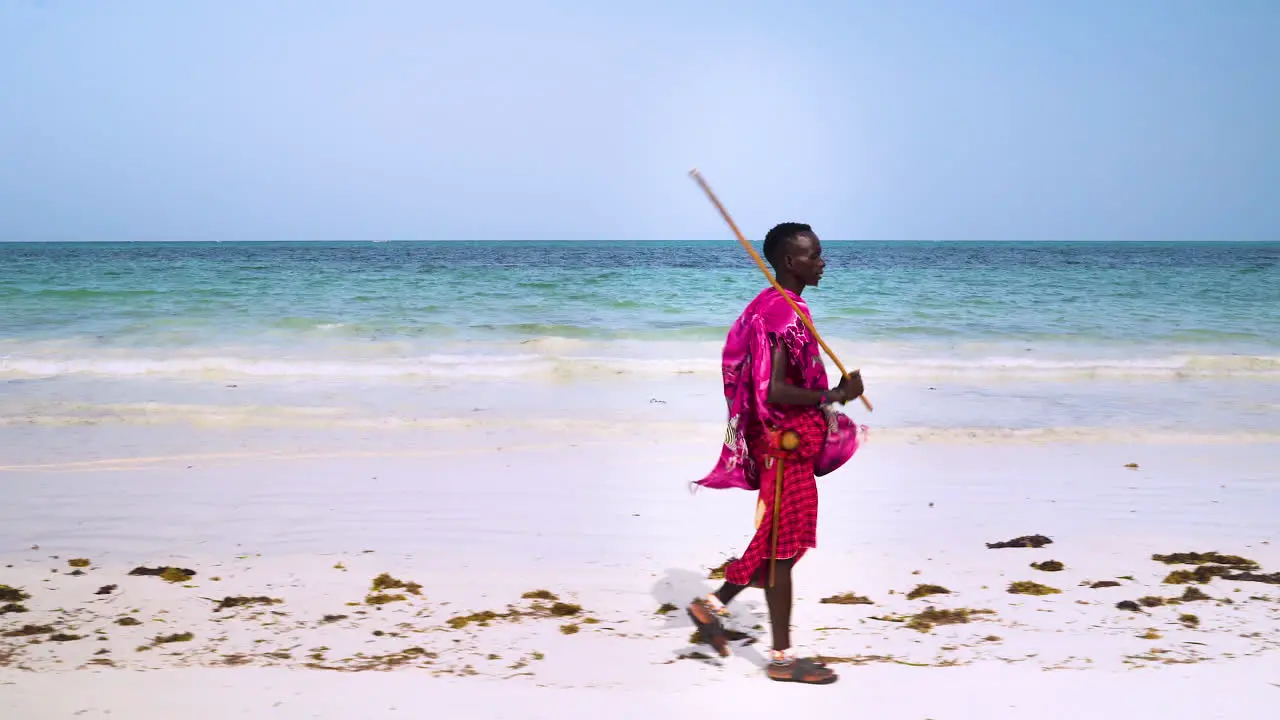 Maasai man in pink clothes walking on sand beach holding wooden stick