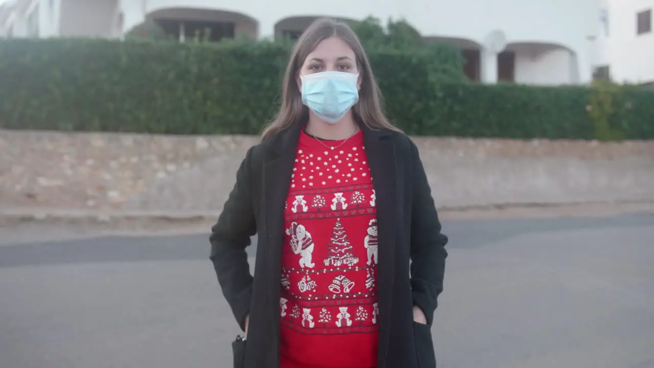 Young adult wearing a Christmas sweater putting on a surgical mask in the street and looking at the camera