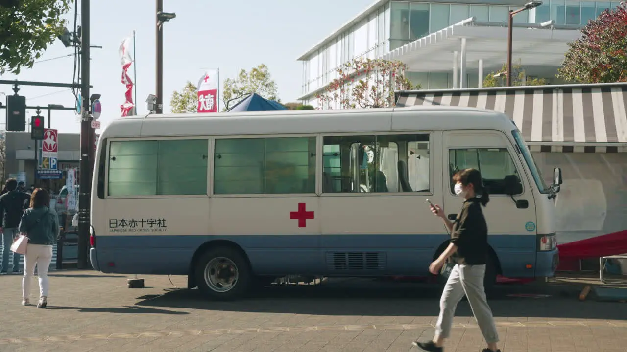 People In Face Mask Passing By Bloodmobile Parked On The Red Cross Blood Donation Station In The Street Of Tokyo During The COVID-19 Outbreak In Tokyo Japan