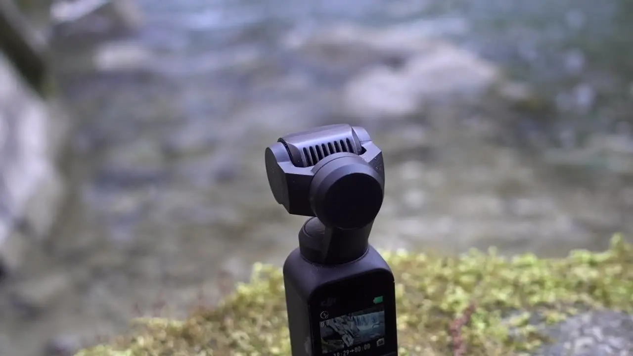 A dji osmo pocket camera is placed on a moss support at the edge of a river in the swiss alps