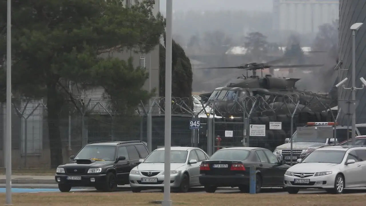 A military helicopter taxis across an airfield Civilian cars parked outside airport security fencing