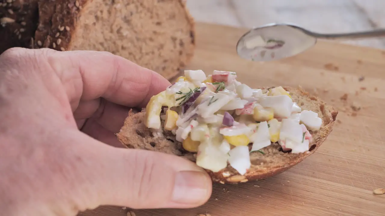 A man puts a vegetable salad on a piece of bread and adds a sprig of dill on a wooden cutting board