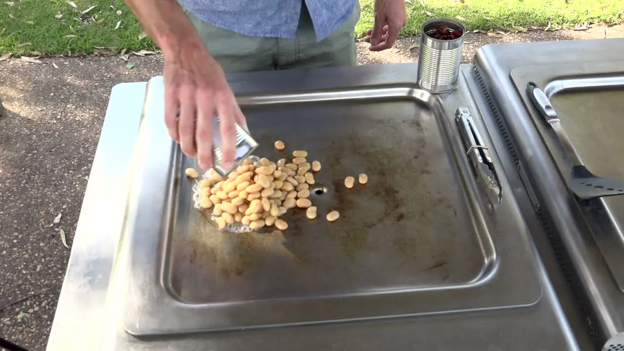 Person pours can of beans onto outdoor cooking hot plate medium shot