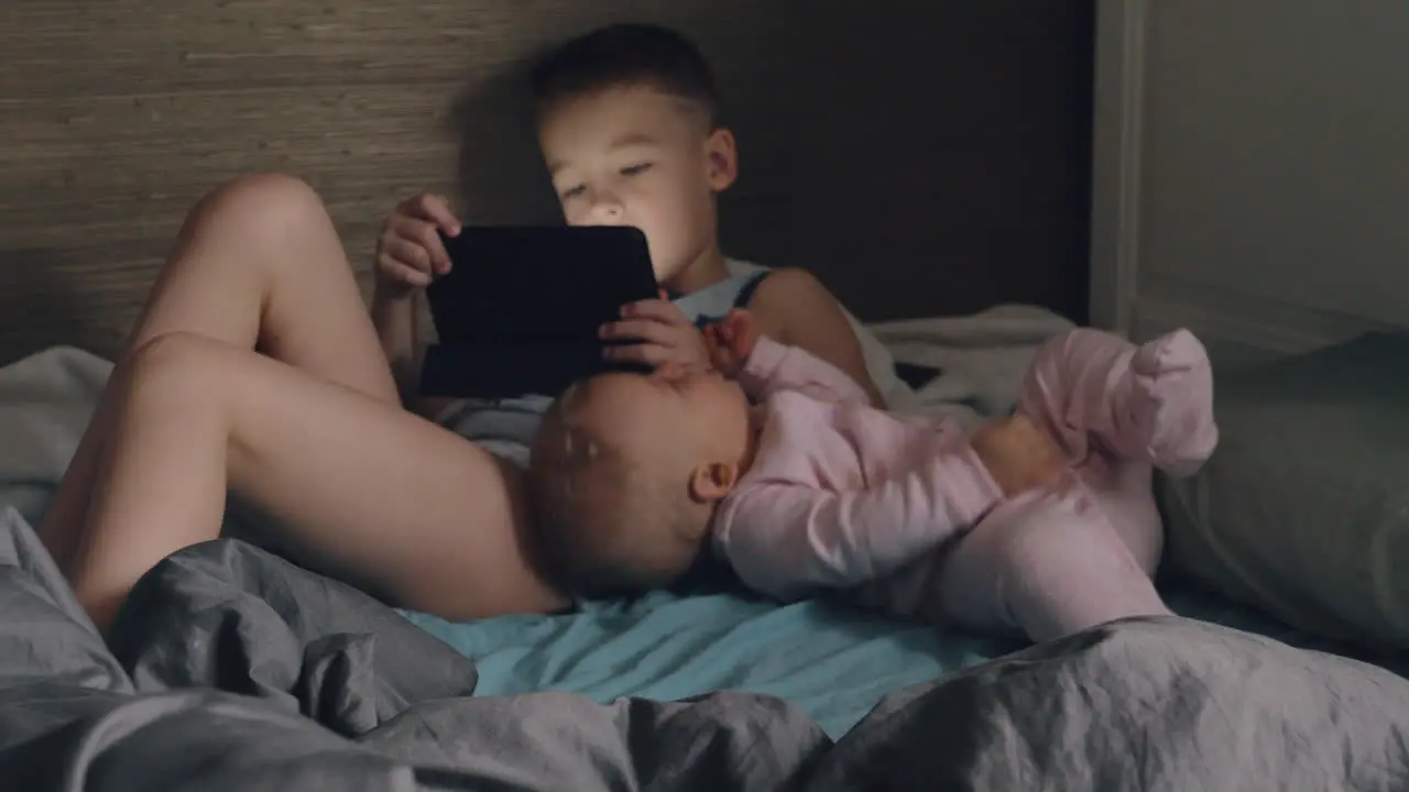 A boy with a tablet and his baby sister on a messy bed