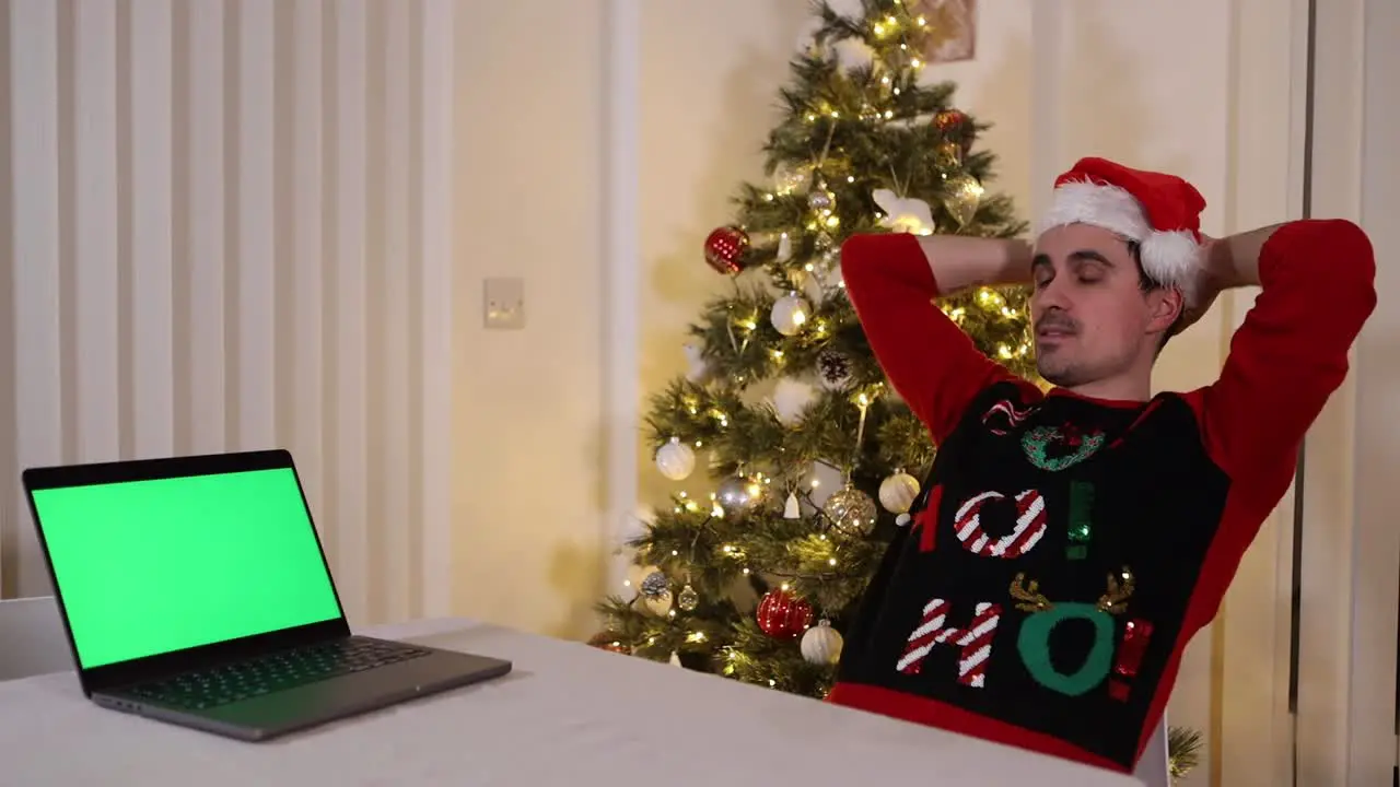 caucasian male wearing Santa hat looking at his laptop with green screen during Christmas holiday while laid back on a comfortable chair in his house