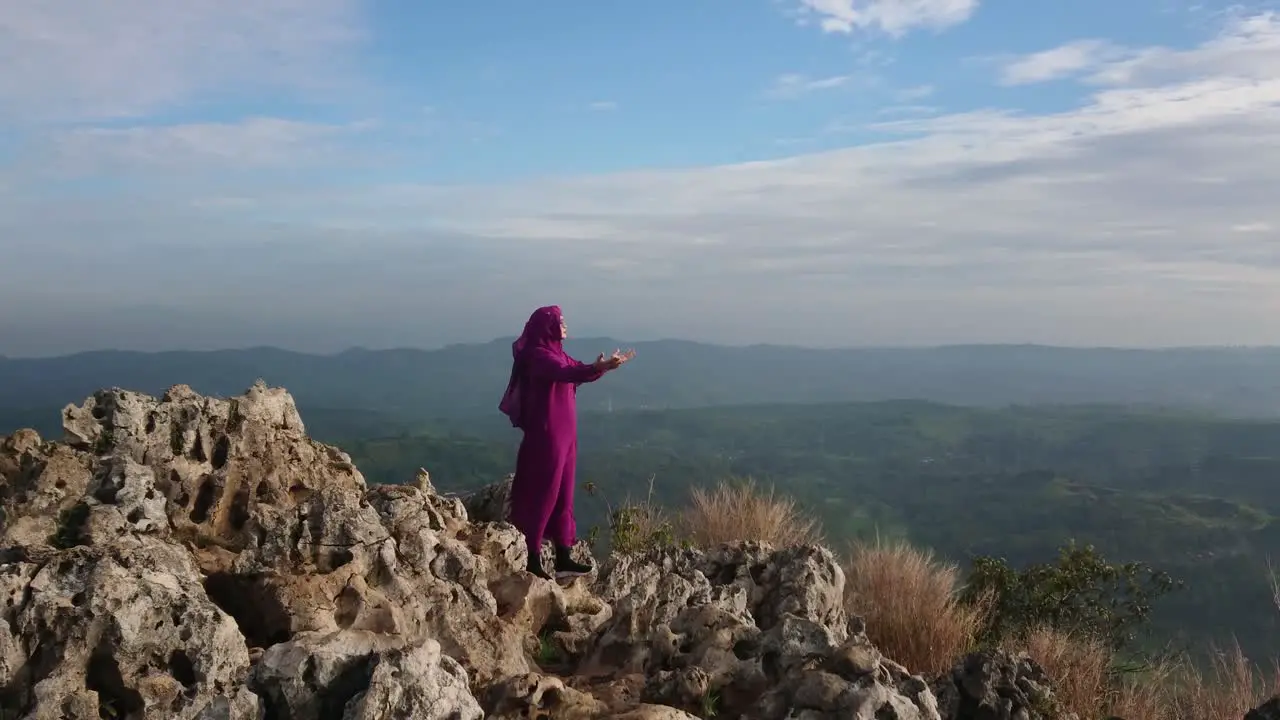 Islamic woman in purple burqa holding hands out in prayer to god on mountain