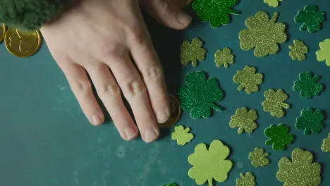 Overhead Studio Shot Of Hand Collecting Gold Coins Next To Shamrock Shapes On Background To Celebrate St Patricks Day