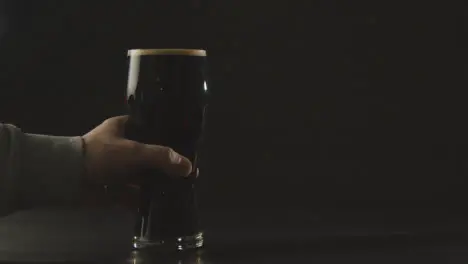 Person Picking Up Pint Of Irish Stout In Glass Against Black Studio Background To Celebrate St Patricks Day