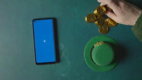 Overhead Studio Shot Of Green Leprechaun Top Hat With Gold Coins And Blue Screen Mobile Phone To Celebrate St Patricks Day 2