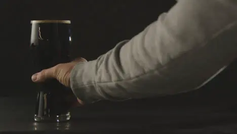 Person Picking Up Pint Of Irish Stout In Glass Against Black Studio Background To Celebrate St Patricks Day 1