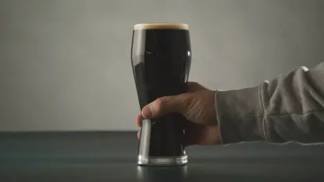 Person Picking Up Pint Of Irish Stout In Glass Against Studio Background To Celebrate St Patricks Day