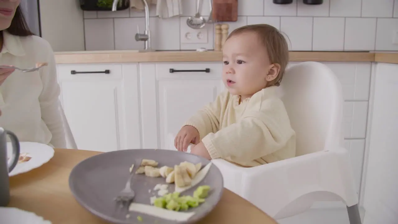 Cute Little Girl Sitting In High Chair In The Kitchen While Her Mother Feeding Her 2