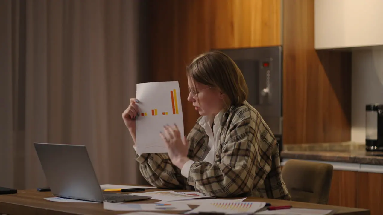 Home office A young woman with glasses shows a graph to a laptop camera while sitting at home in the kitchen conducting an online report Video call and conference with demonstration of documents