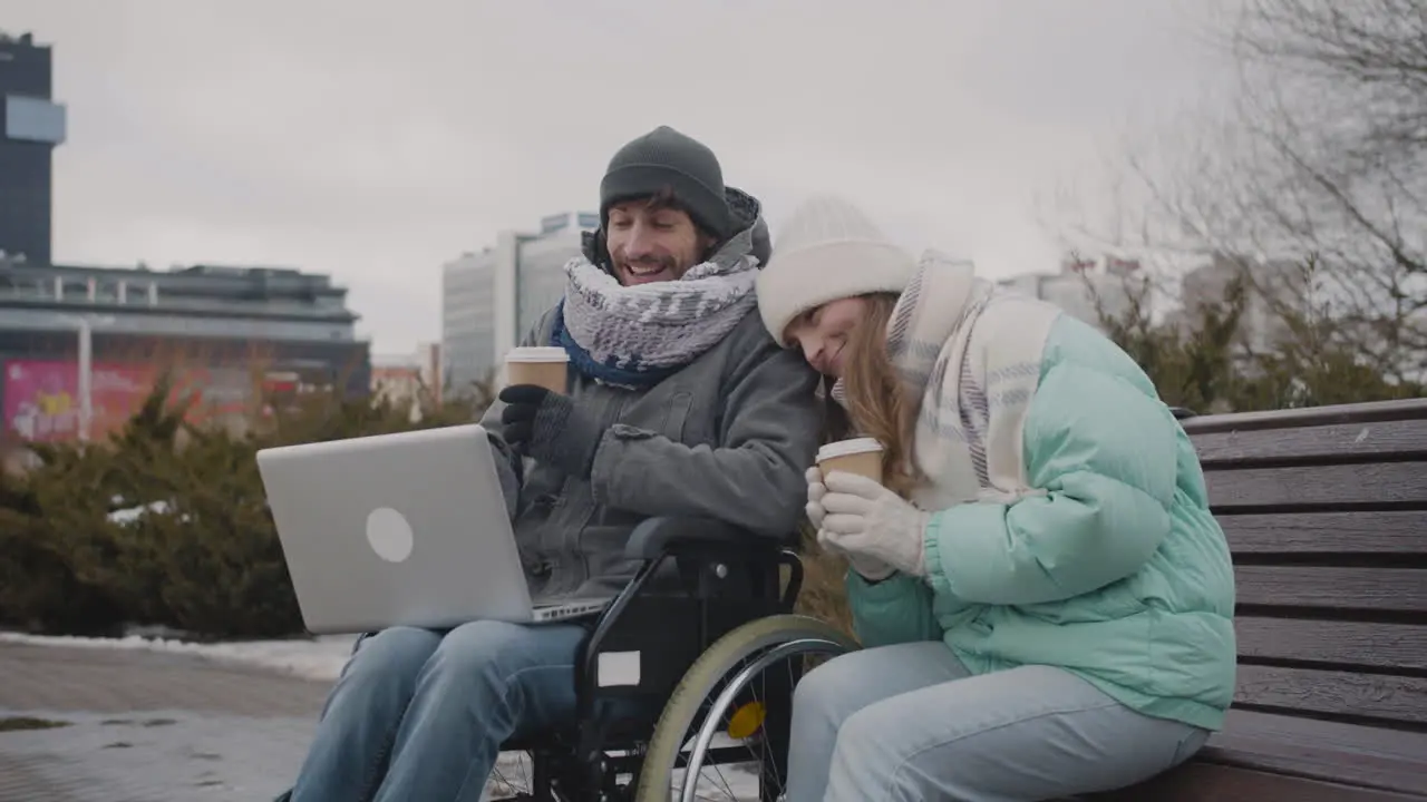 Disabled Man In Wheelchair And His Friend Watching Something Funny On Laptop Computer And Laughing Together At Urban Park In Winter 1