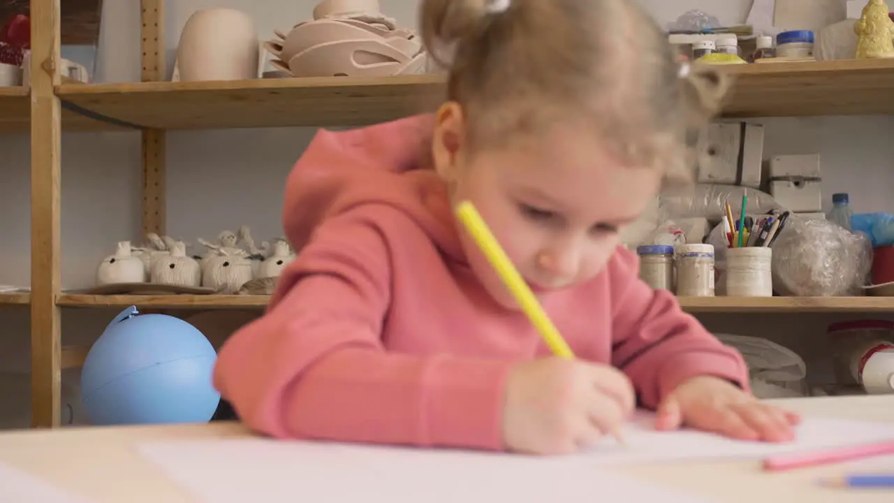 Blonde Little Girl Drawing On A Paper Sitting At A Table In A Craft Workshop 1