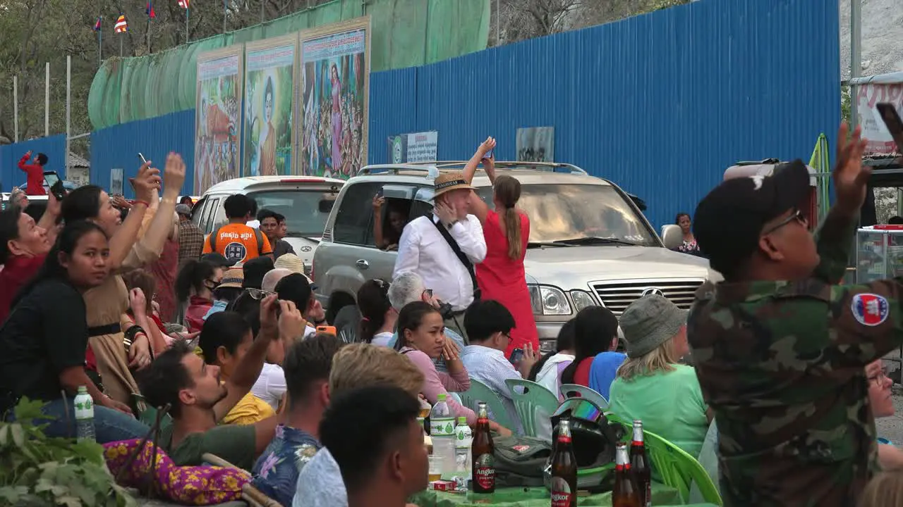 Medium Exterior Shot of Tourists Looking Up and Filming With Their Phones While Others are Sitting With Refreshment as Traffic is Moving Slowly in Front of Them