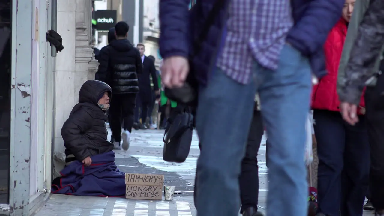 People walk past a homeless person sat on Oxford Street begging for money with a cardboard sign that reads I am very hungry God bless”