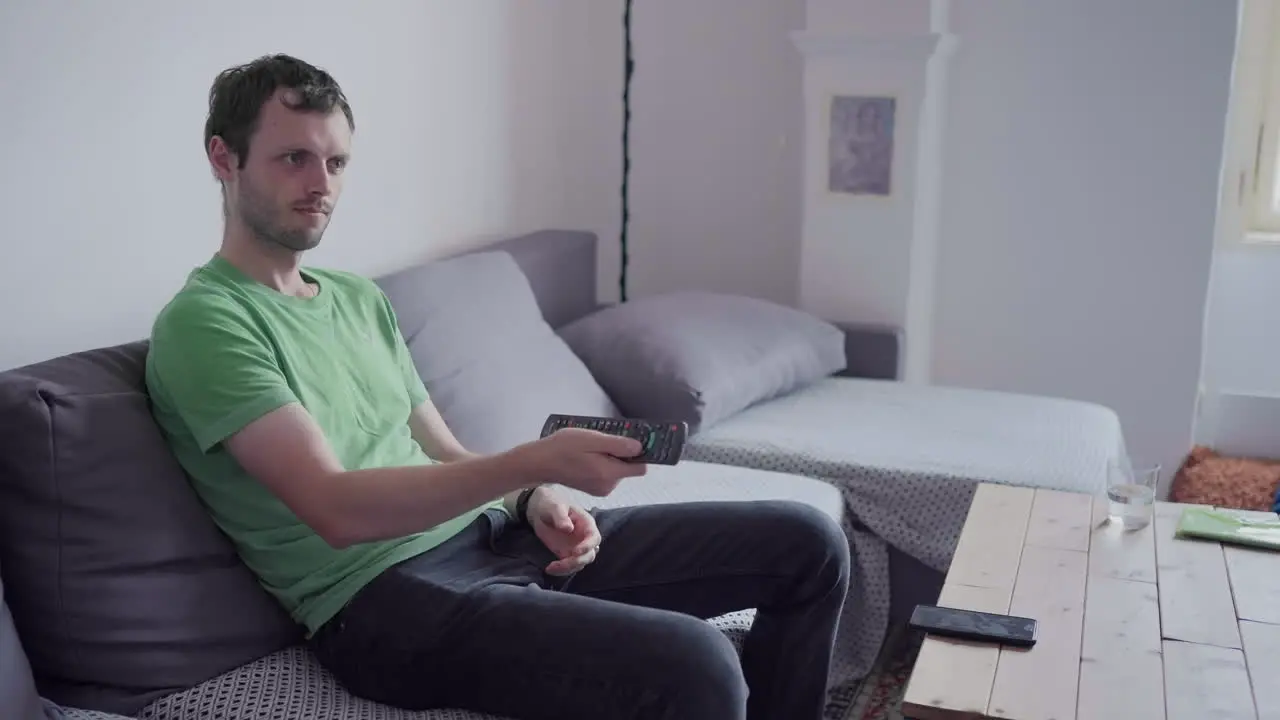 Young man with dark hair watching tv and changing the channels with remote control