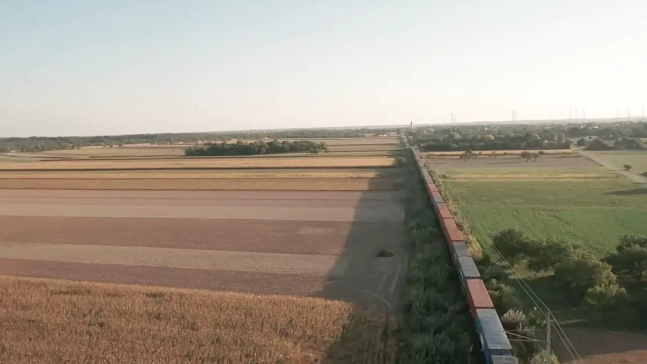 Cinematic descending aerial drone captures a laden cargo train traversing vast golden-lit cornfields while the drone glides overhead unveiling the serene rural scenery