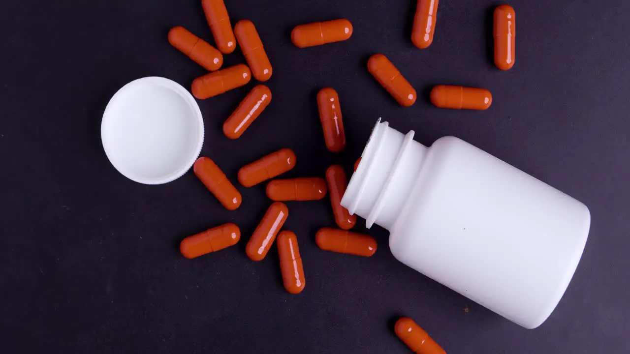 Birdyeye shot of a rotating white tablet box with red tablets distributed in slow motion on black background