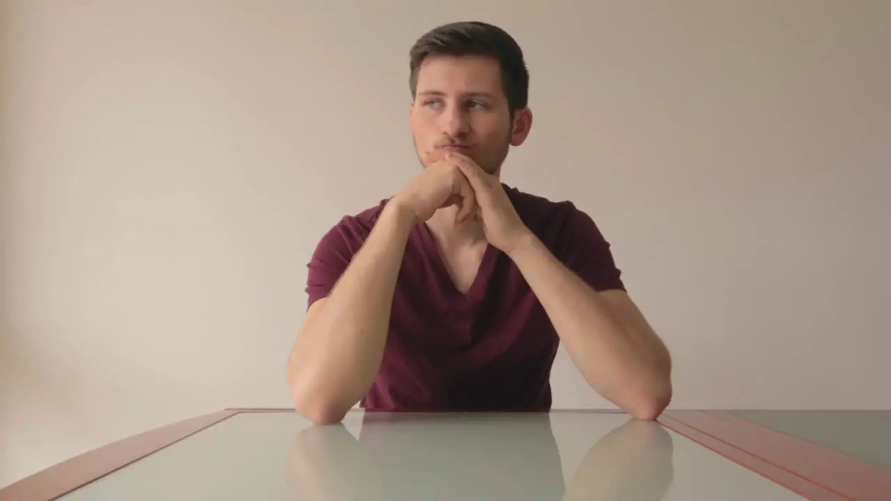 Young man sitting at table being indecisive about a choice and thinking hard medium close-up shot facing camera indoors with natural light