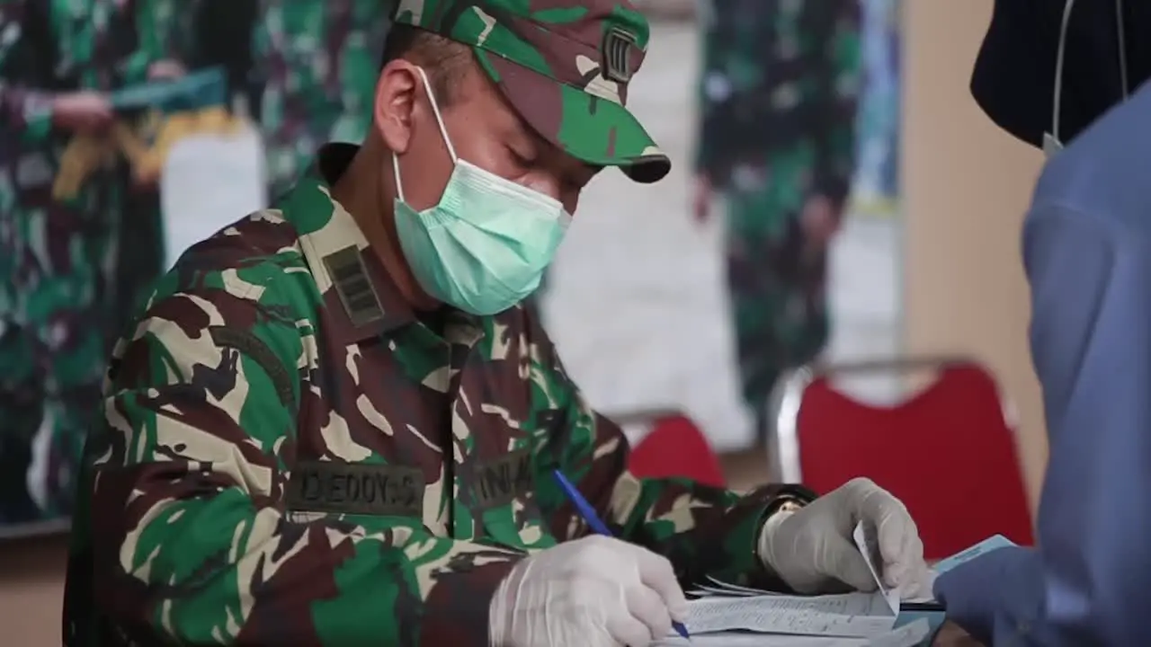 A male doctor from the Indonesian National Armed Forces is writing or registering patients assisted by a female nurse at the registration desk in front of the hospital