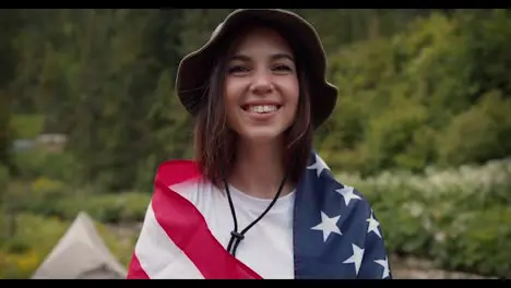 A happy brunette girl in a green hat wrapped herself in the flag of the United States of America smiles and looks at the camera against the background of a green forest and a mountain river