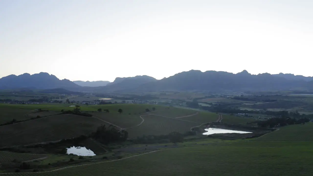 Aerial drone over green vineyards and ponds with blue mountains and farmhouse shed in background before sunrise Stellenbosch