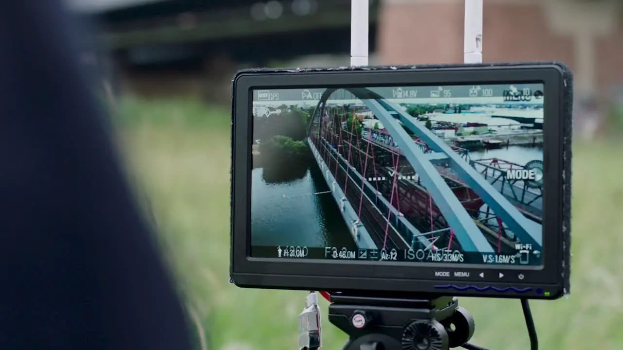 Handheld camera view of monitor attached to tripod showing red arched bridge over a river trees and buildings in background with settings visible on display