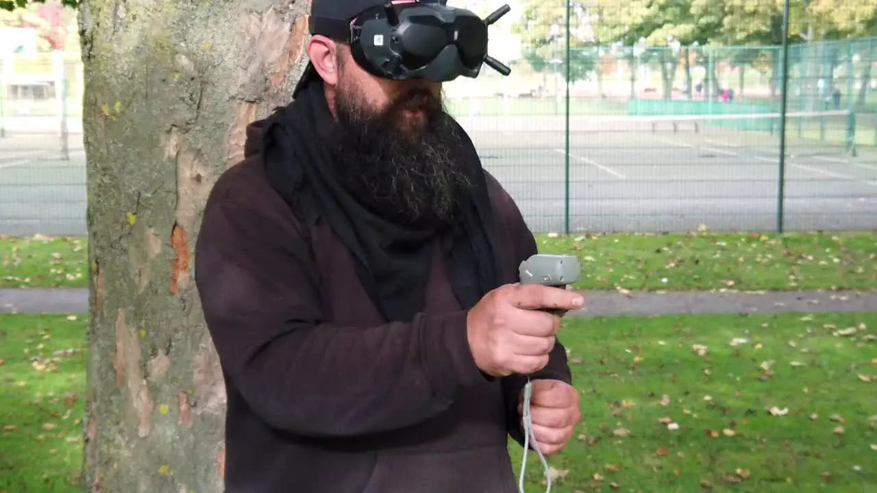 Bearded male wearing augmented reality FPV goggles controlling drone in park with motion joystick