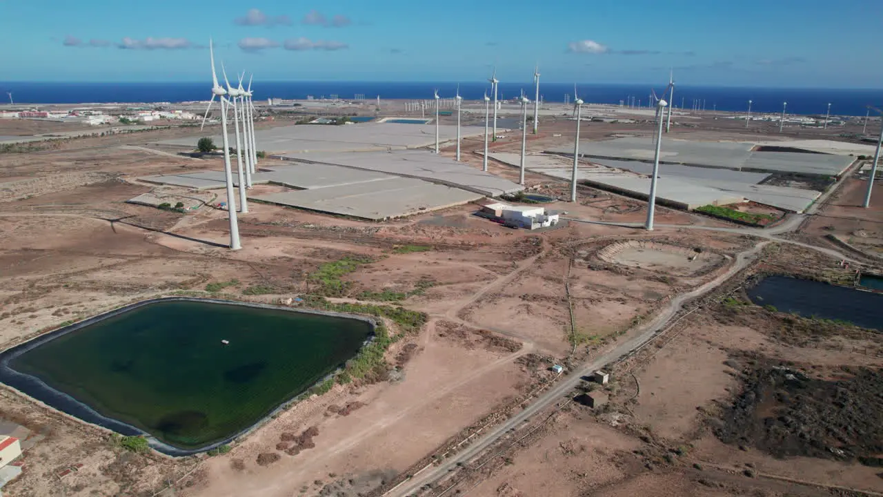 Aerial view in orbit over a field of wind turbines and a pond of water in a desert landscape and where the ocean can be seen