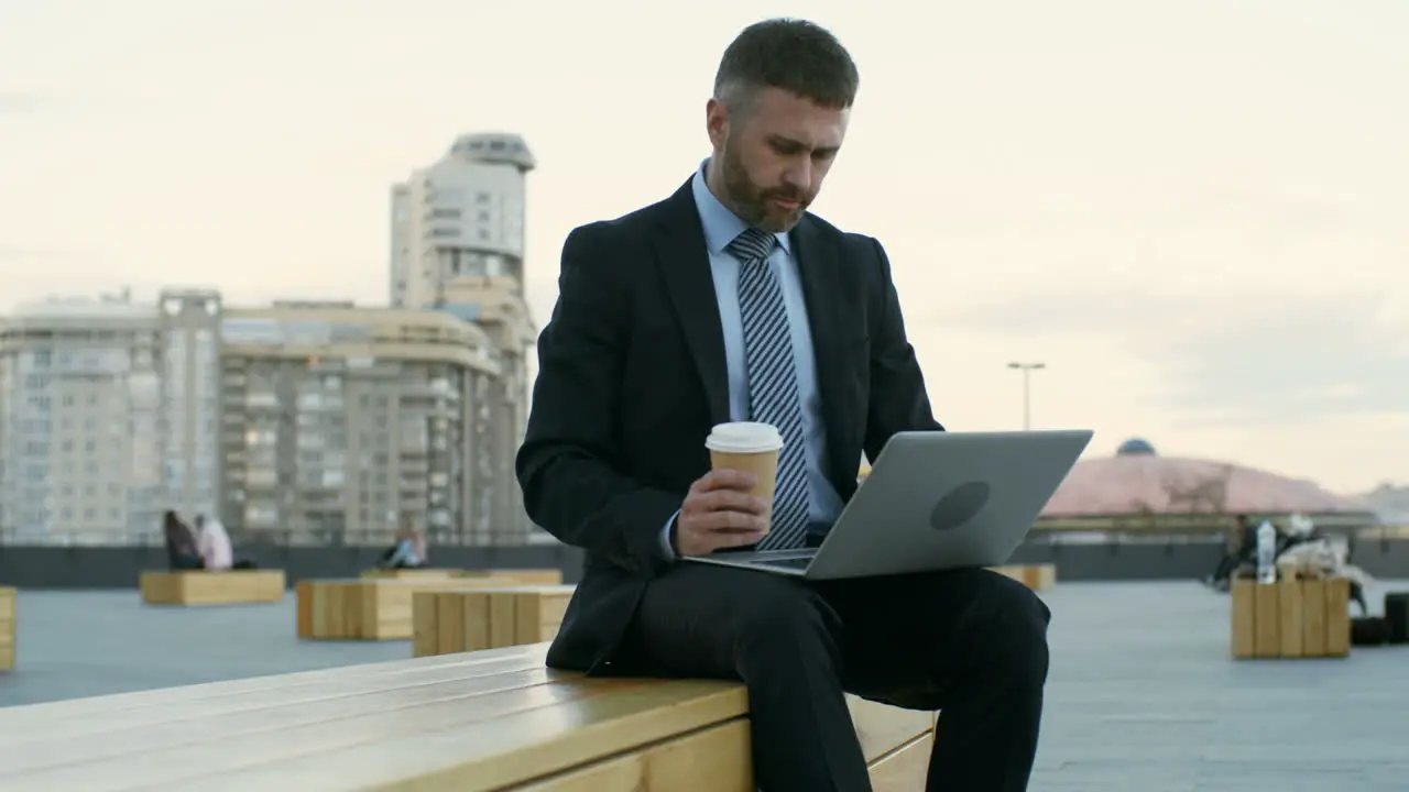 Businessman Sitting Outdoors On Wooden Bench With Laptop On His Knees Typing On Keyboard And Drinking Coffee