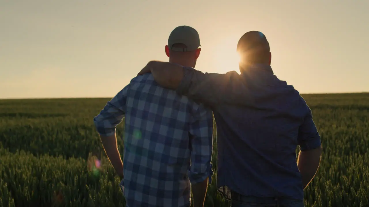 The farmer hugs his adult son and looks forward together to the wheat field where the sun is setting Family business and traditions concept