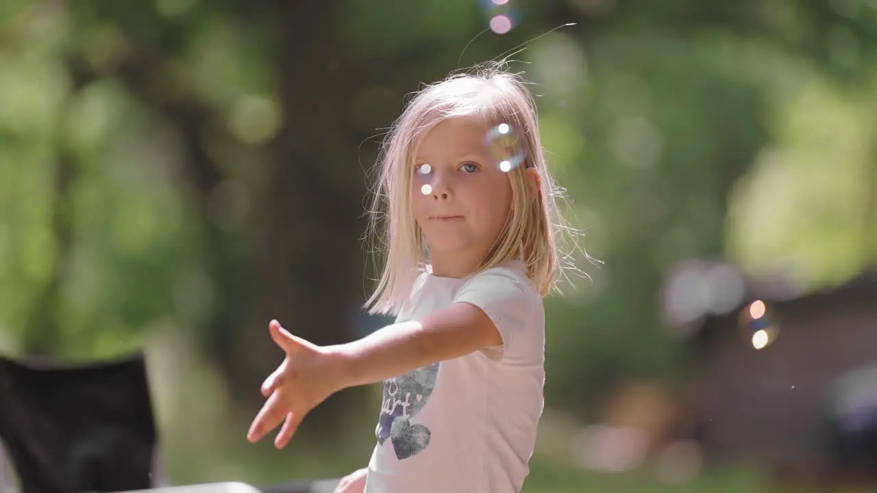 A bright-eyed blonde girl enjoys a sunny day outdoors as she delicately blows soap bubbles that shimmer and float around her