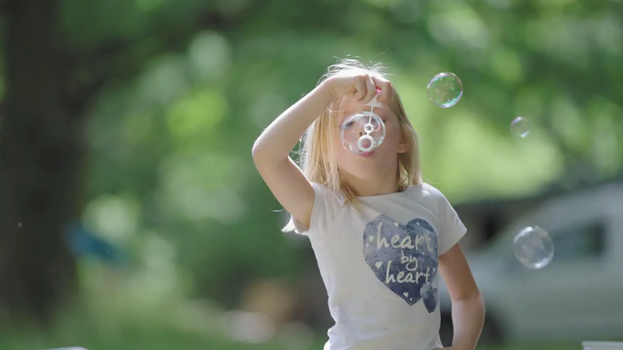 A blonde-haired little girl embraces the simple joy of a sunny day blowing soap bubbles outside