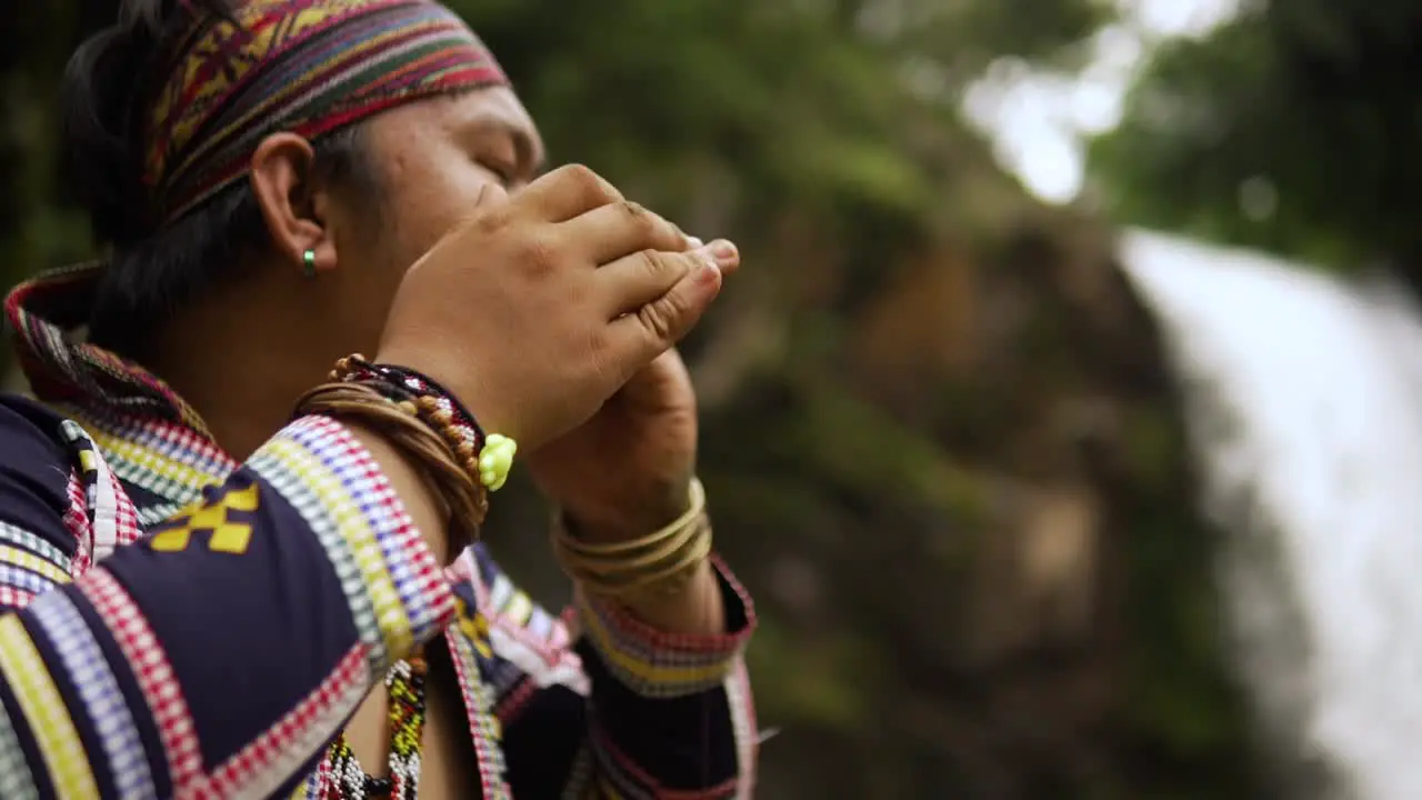 orbit shot of an Indigenous person playing Kubing tribal instrument behind is a waterfall