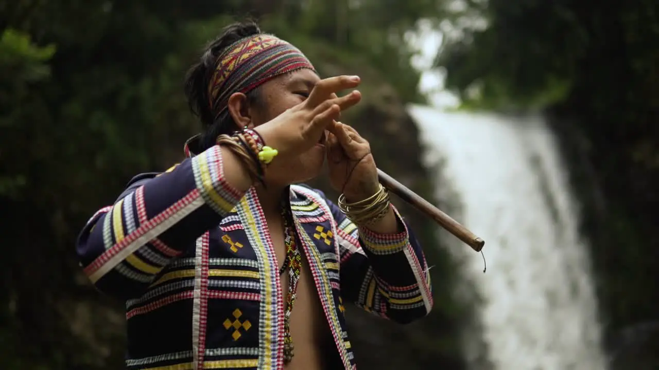 orbit shot of Indigenous person playing Kubing tribal instrument behind is a waterfall while standing