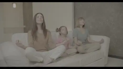 Two mature women and a little girl are sitting on the sofa in a yoga pose and meditating