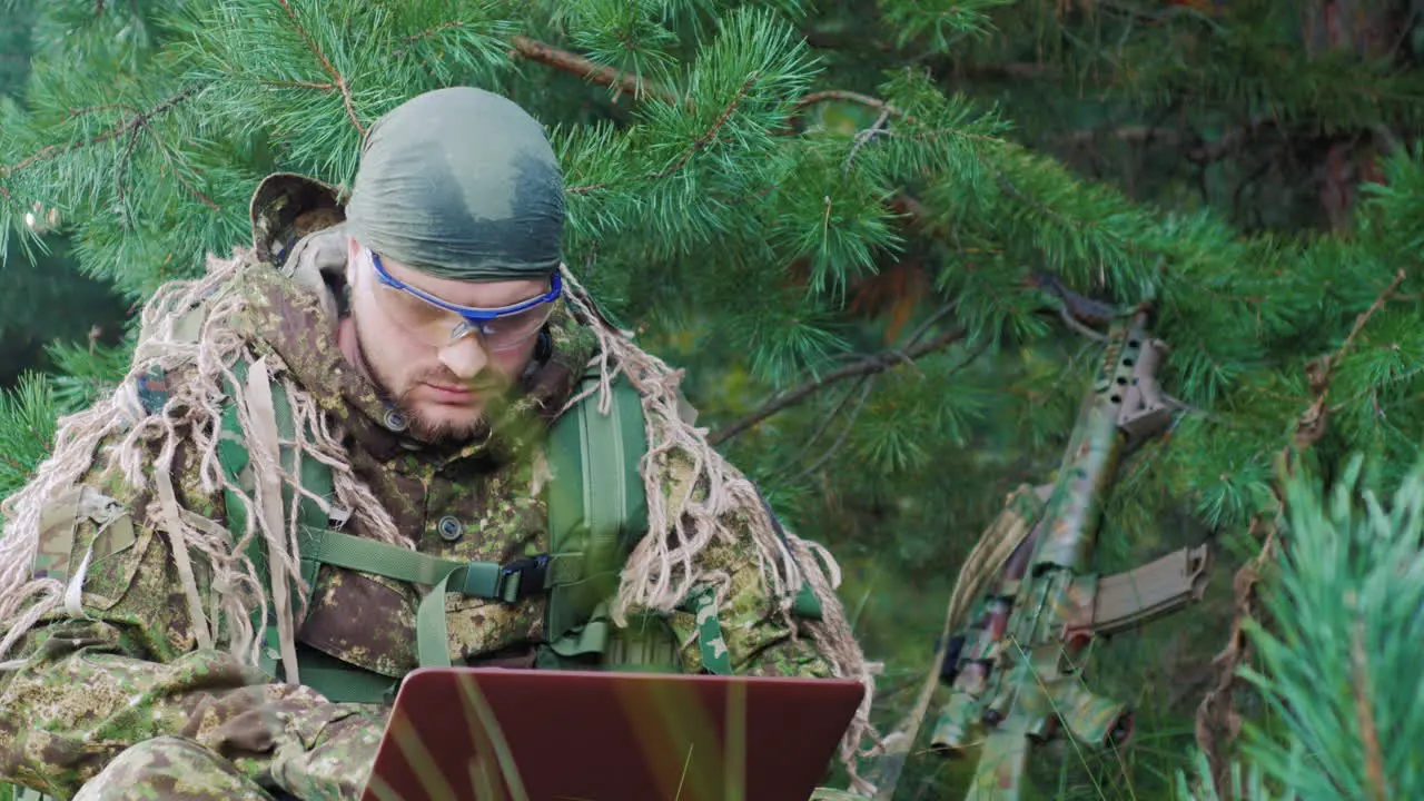 Portrait Of Military Man Sitting In The Woods Dressed In Camouflage It Uses Laptop