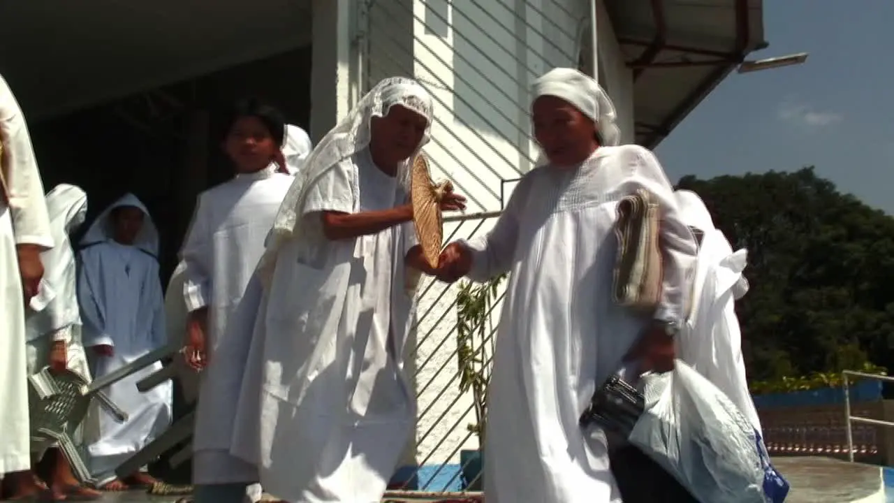 A very old woman wearing white dress is having difficulty in going down the stairs from a church Another women are helping her