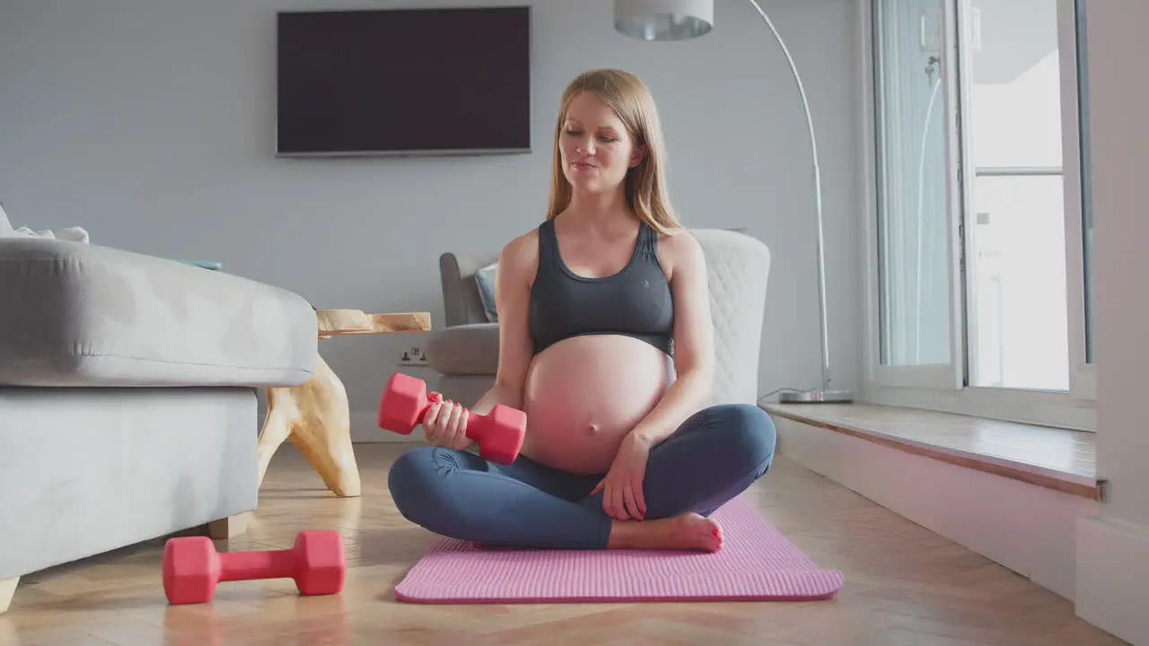 Pregnant Woman Wearing Fitness Clothing On Mat At Home Exercising With Weights