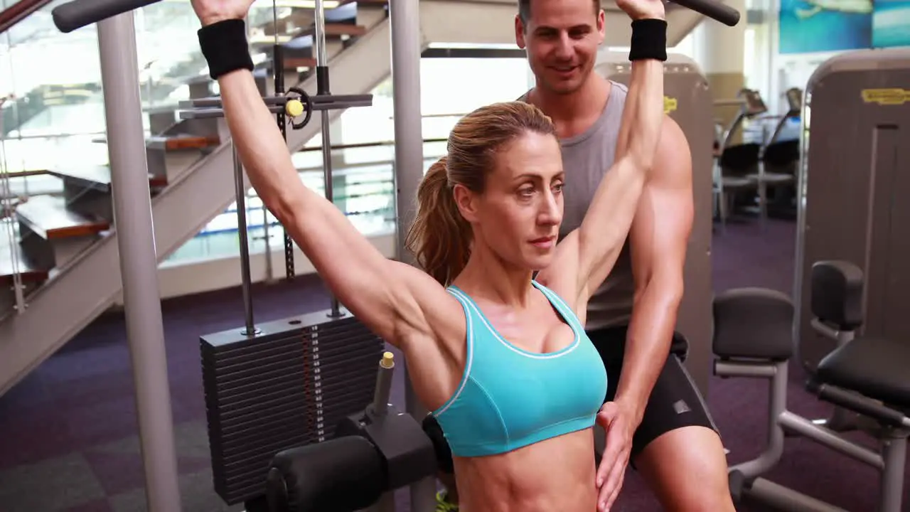 Fit woman using the weights machine for her arms while trainer supervises