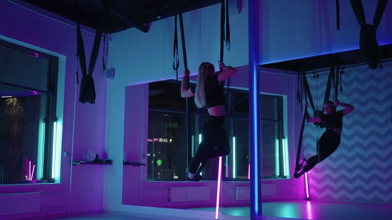 A woman does sports yoga in the air on a hammock in neon light performing graceful exercises and movements from stretching and dancing in slow motion