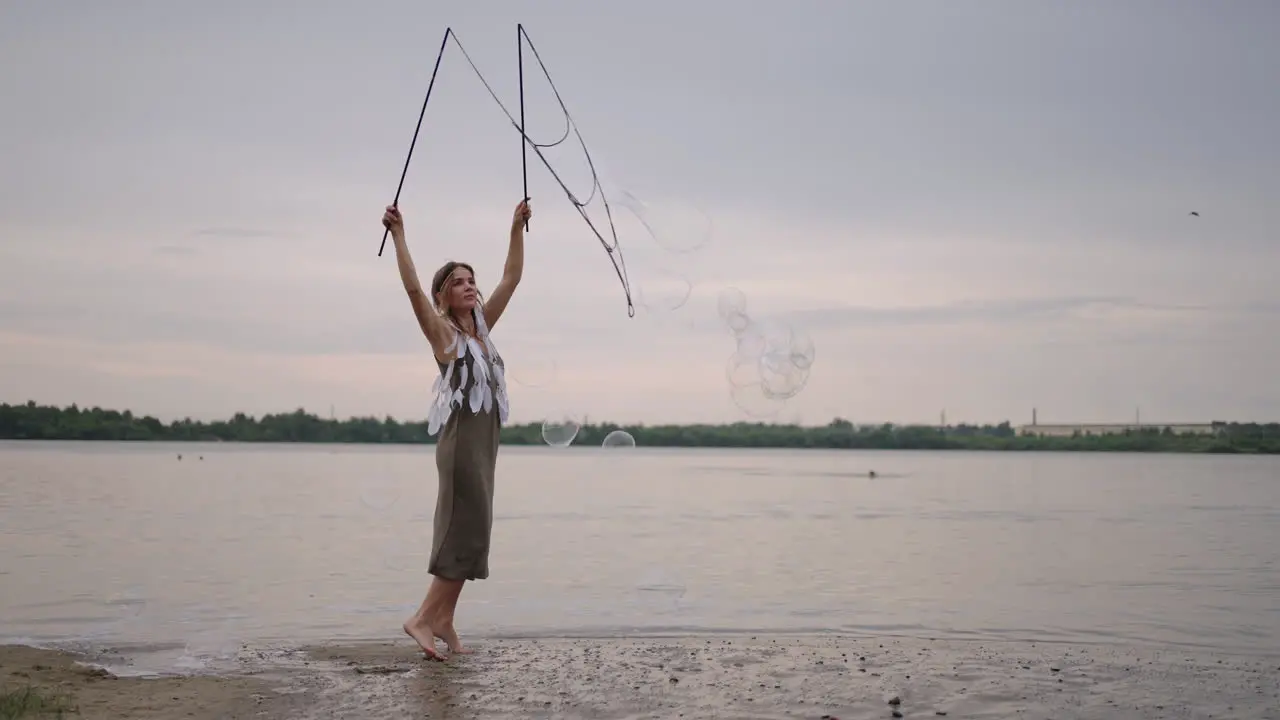 A young hippie woman in a dress and with feathers on her head makes huge soap bubbles at sunset on the shore of a lake in slow motion